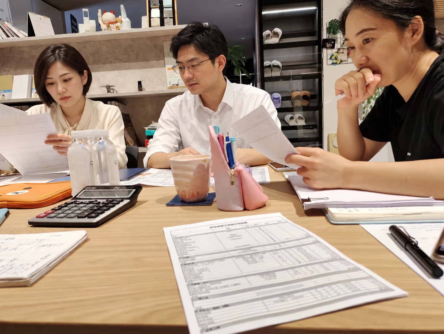 三人围桌阅读资料并交流,桌面摆放图纸、计算器和记录用品
